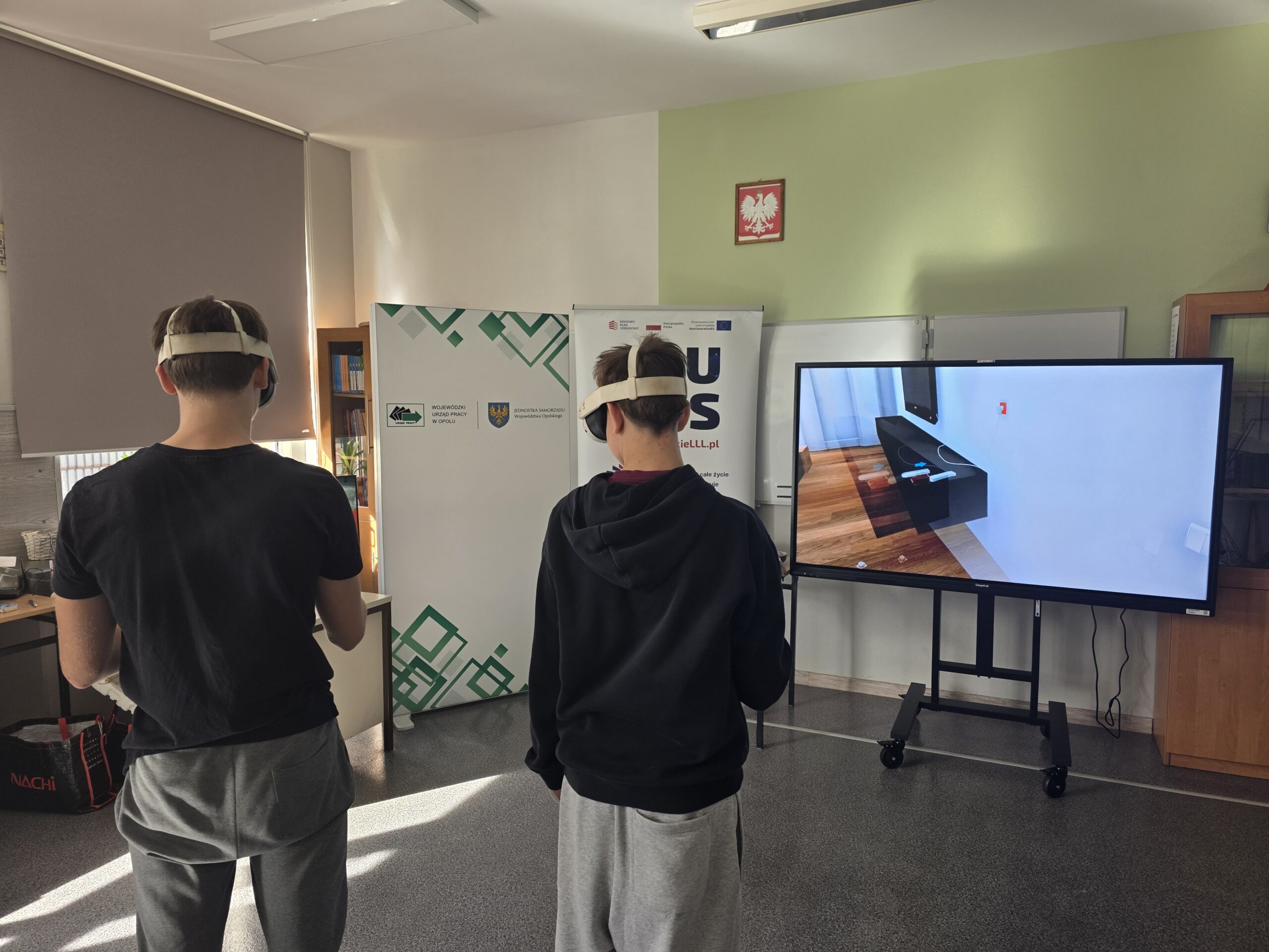W sali lekcyjnej stoją dwaj uczniowie w goglach VR, wpatrując się w duży ekran telewizyjny, na którym wyświetlana jest wirtualna symulacja aranżacji wnętrz.