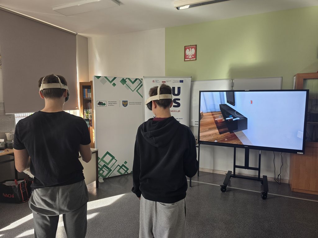 Chłopak stoi tyłem. Ma na głowie gogle VR. Przed nim stoją dwa roll upy z informacją o EduBusie oraz Wojewódzkim Urzędzie Pracy w Opolu. Po prawej stronie stoi duża tablica multimedialna.