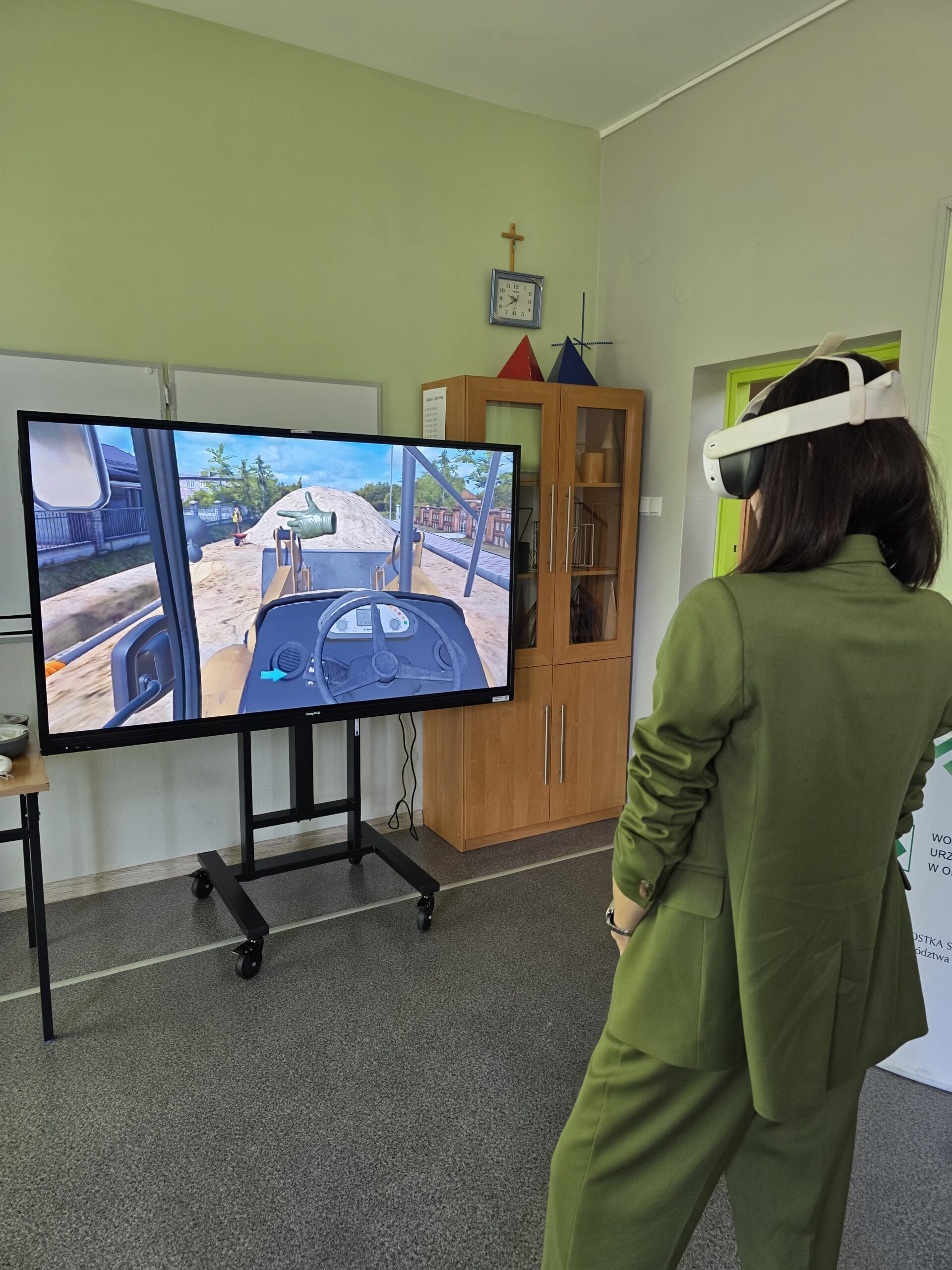 Osoba nosząca gogle VR siedzi przed dużym ekranem, na którym wyświetlana jest symulacja pracy maszyny budowlanej. Ekran przedstawia widok z wnętrza kabiny spycharki pracującej na placu budowy.