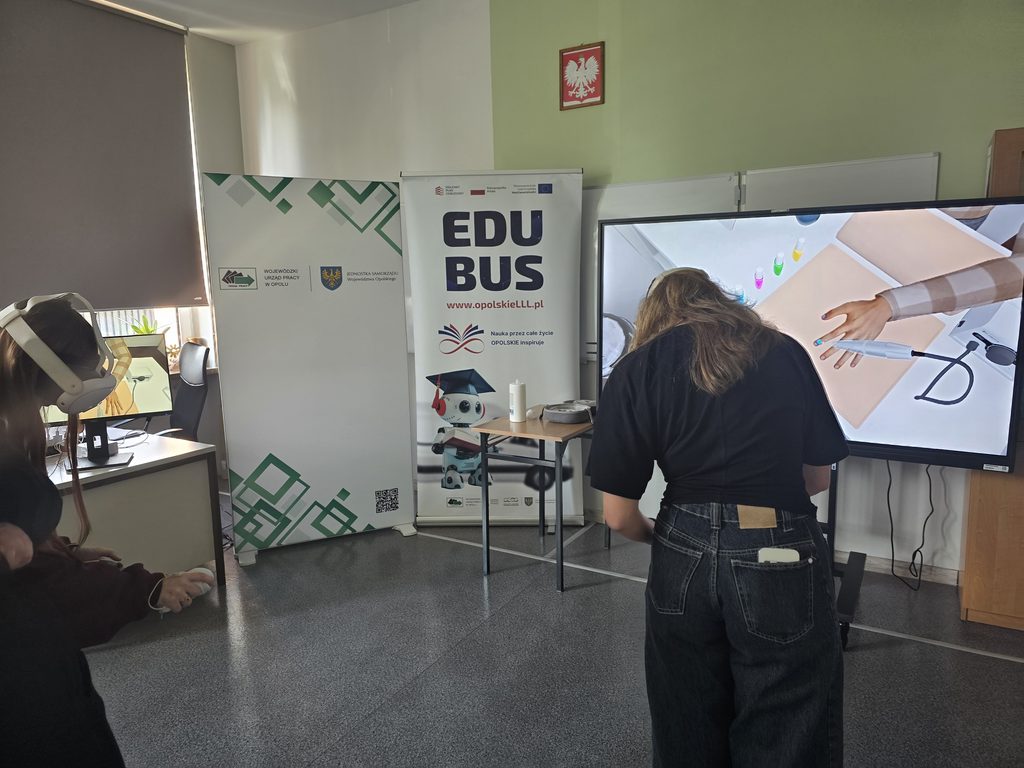 Dziewczyna stoi tyłem. Ma na głowie gogle VR. Przed nią stoją dwa roll upy z informacją o EduBusie oraz Wojewódzkim Urzędzie Pracy w Opolu. Po prawej stronie stoi duża tablica multimedialna.