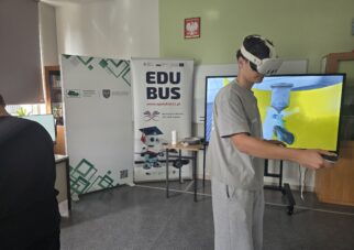 Chłopak stoi bokiem do odbiorcy zdjęcia. Ma na głowie gogle VR. Za nim stoją dwa roll upy z informacją o EduBusie oraz Wojewódzkim Urzędzie Pracy w Opolu. Po prawej stronie stoi duża tablica multimedialna.