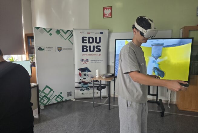 Chłopak stoi bokiem do odbiorcy zdjęcia. Ma na głowie gogle VR. Za nim stoją dwa roll upy z informacją o EduBusie oraz Wojewódzkim Urzędzie Pracy w Opolu. Po prawej stronie stoi duża tablica multimedialna.