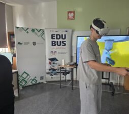 Młody mężczyzna w goglach VR trzyma kontrolery, patrząc na ekran, na którym wyświetla się trójwymiarowa symulacja przemysłowa. Za nim wisi baner marki EduBus z ilustracją robota.