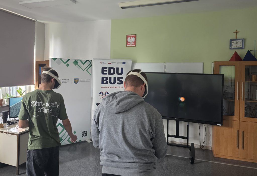 Dwóch chłopców stoi tyłem. Mają na głowie gogle VR. Przed nimi stoją dwa roll upy z informacją o EduBusie oraz Wojewódzkim Urzędzie Pracy w Opolu. Po prawej stronie stoi duża tablica multimedialna.