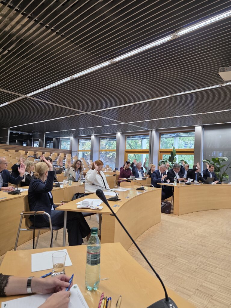 Uczestnicy spotkania w sali konferencyjnej podnoszą ręce do góry podczas głosowania. Osoby siedzą przy długich, łukowatych stołach z mikrofonami, dokumentami i napojami. Na pierwszym planie widać dłoń trzymającą długopis oraz butelkę wody. Perspektywa ujęcia ukazuje głębię sali z podwyższonymi rzędami siedzeń w tle.