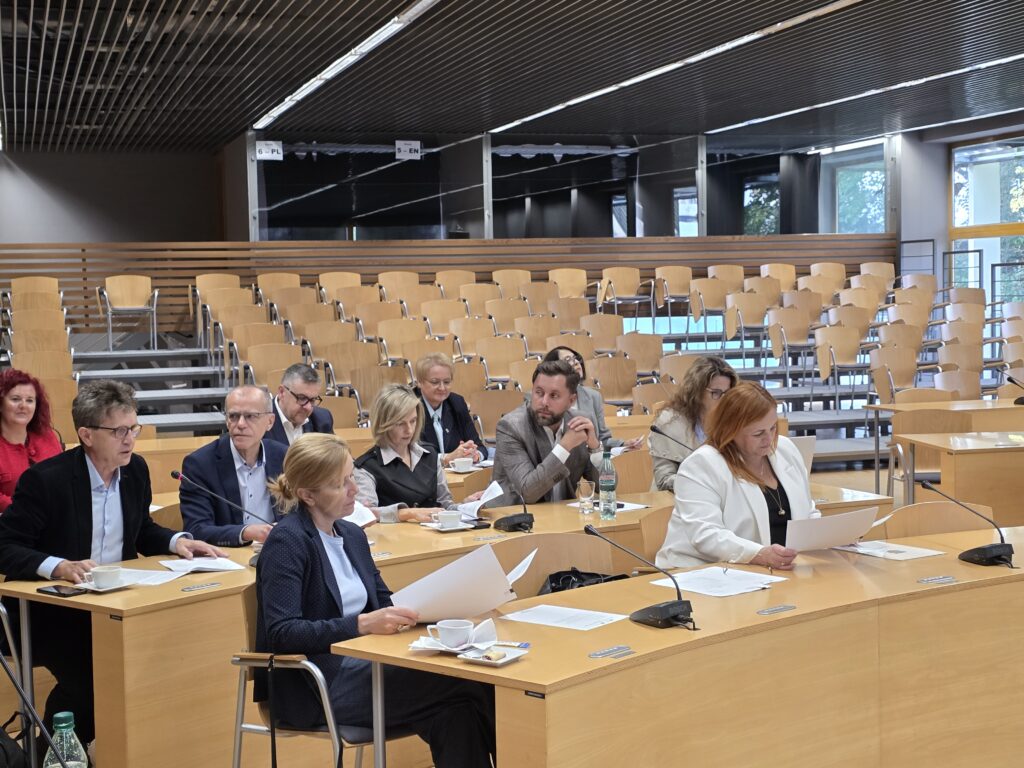Szeroki kadr przedstawiający grupę osób siedzących przy łukowato ułożonych stołach w jasnej sali obrad. Uczestnicy spotkania są elegancko ubrani, czytają materiały prasowe lub słuchają wypowiedzi. Przed nimi na stołach znajdują się mikrofony, filiżanki z napojami oraz butelki wody. Tło stanowi pusta widownia z drewnianymi krzesłami.