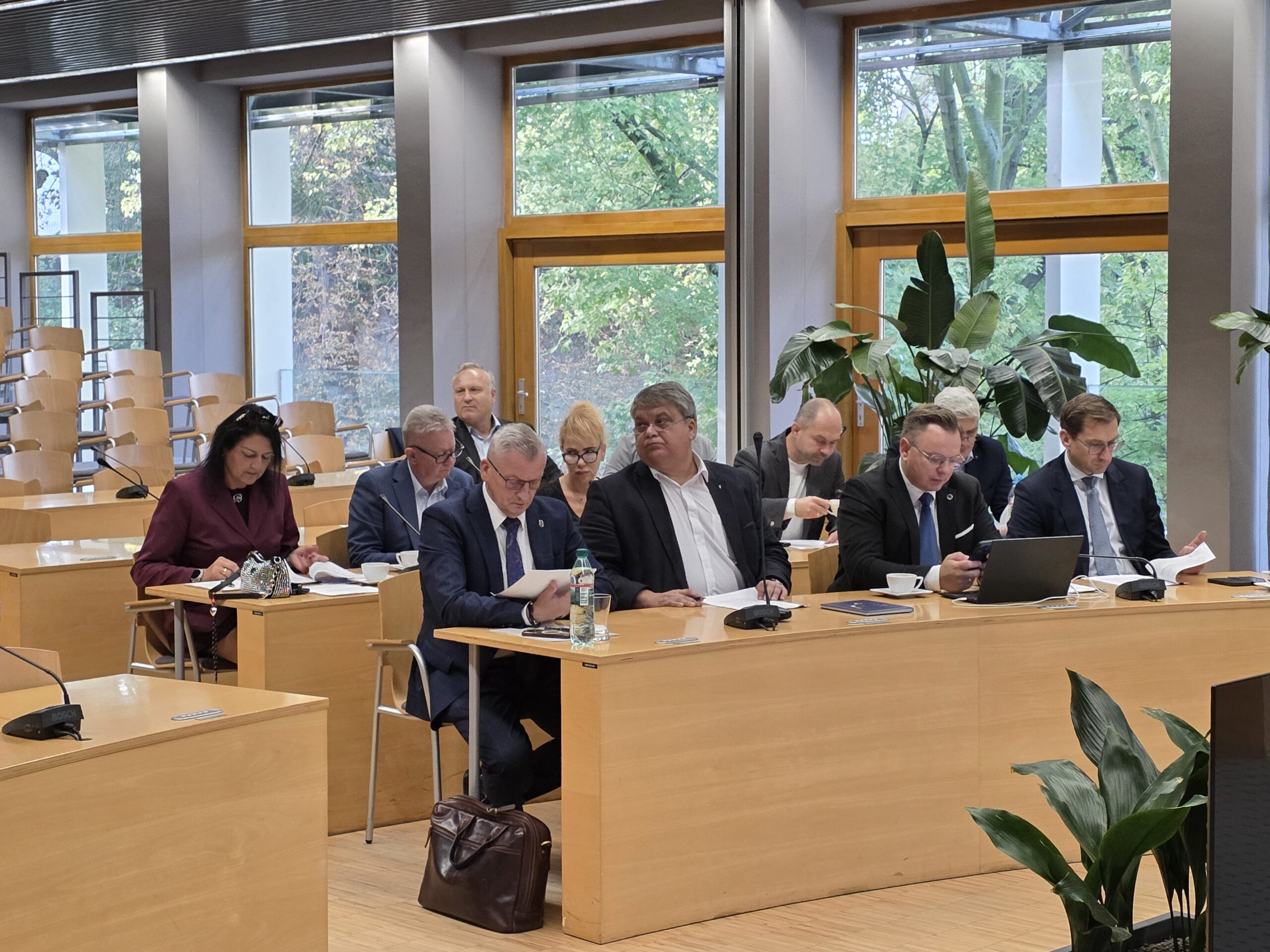 Uczestnicy spotkania w sali konferencyjnej siedzą przy stołach ustawionych tyłem do dużych okien. Część osób analizuje dokumenty papierowe, a jedna z nich korzysta z otwartego laptopa. Przez przeszklone ściany w tle widać zielone drzewa oraz rośliny doniczkowe wewnątrz pomieszczenia