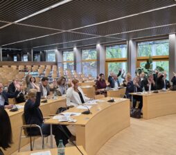 Głosowanie podczas oficjalnego spotkania w dużej sali audytoryjnej. Większość zgromadzonych przy stołach osób podnosi prawą rękę w górę. Stoły ułożone są w półokręgu, a cała przestrzeń jest dobrze oświetlona
