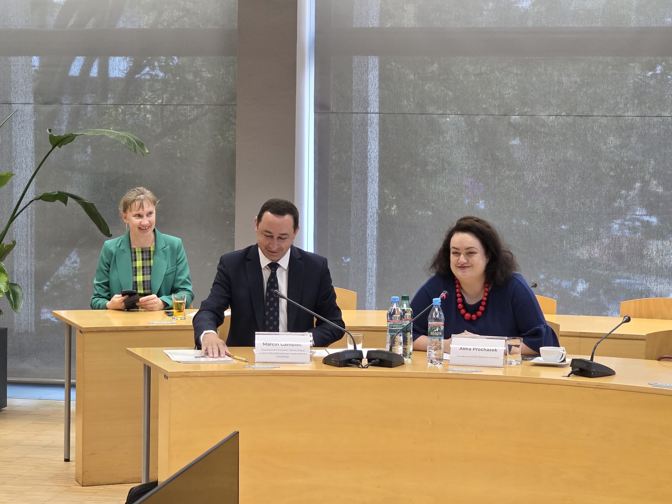 Trzy osoby siedzące przy stole prezydialnym na tle osłoniętych roletami okien. Pośrodku znajduje się mężczyzna w ciemnym garniturze, a po jego prawej stronie kobieta w ciemnoniebieskiej bluzce z czerwonym naszyjnikiem. Przed nimi na stole leżą tabliczki z imionami i nazwiskami, mikrofony oraz butelki z wodą. W tle po lewej stronie uśmiecha się kobieta w zielonej marynarce