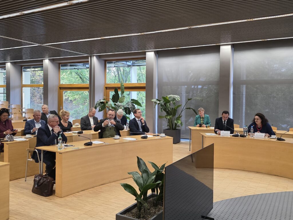 Szerokie ujęcie nowoczesnej, jasnej sali konferencyjnej ułożonej w kształcie podkowy. Przy stołach siedzą uczestnicy obrad, a w centralnej części widoczny jest stół prezydialny oraz duże rośliny doniczkowe. Duże przeszklenia i rolety w tle wpuszczają do wnętrza naturalne światło.