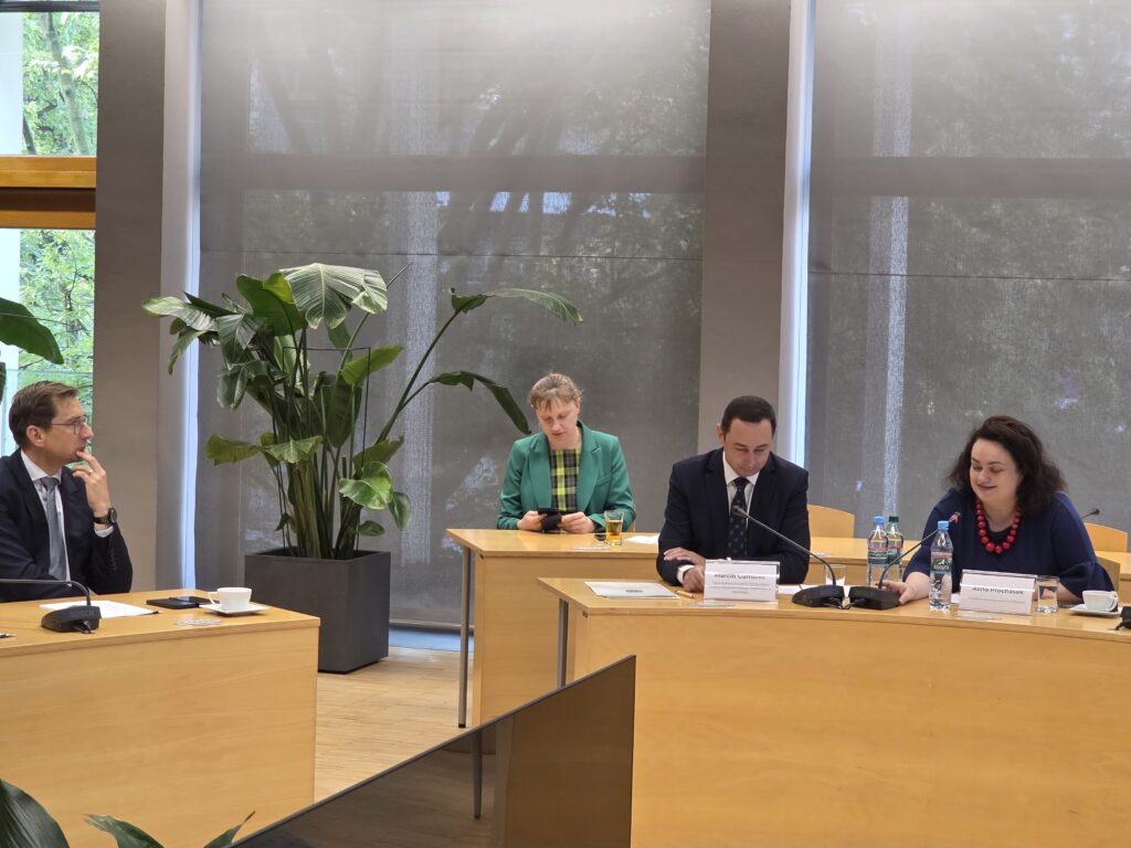 Zbliżenie na stół prezydialny w sali konferencyjnej, przy którym siedzą cztery osoby. Po prawej stronie kobieta w ciemnoniebieskiej bluzce i czerwonym naszyjniku opiera się o stół z tabliczką imienną. Obok niej siedzi mężczyzna w garniturze i krawacie, a dalej kobieta w zielonym żakiecie spoglądająca w telefon. Po lewej stronie widać profil mężczyzny w okularach podpierającego brodę.