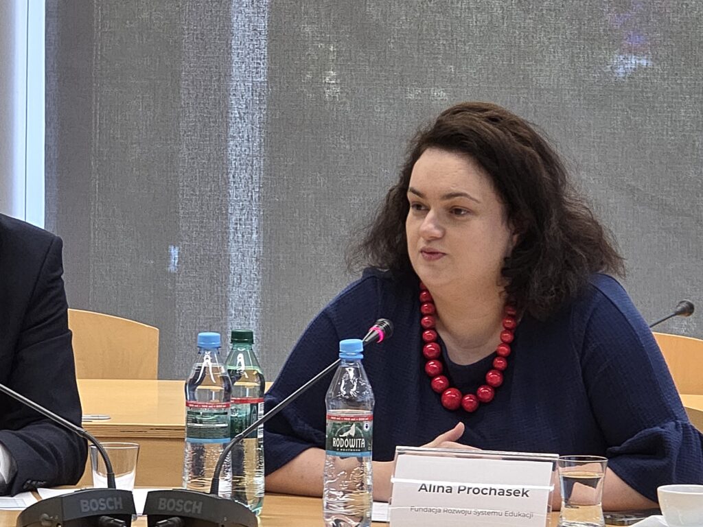 Portret kobiety o ciemnych, kręconych włosach, siedzącej przy stole konferencyjnym. Ma na sobie granatową bluzkę z luźnymi rękawami oraz wyrazisty, czerwony naszyjnik z dużych koralików. Przed nią stoi tabliczka imienna z napisem „Alina Prochasek – Fundacja Rozwoju Systemu Edukacji” oraz butelki z wodą mineralną. Kobieta patrzy w stronę pozostałych uczestników spotkania.