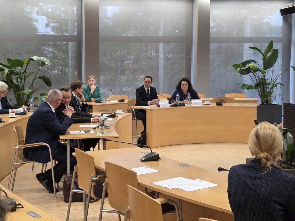 Ujęcie z boku sali na uczestników obradujących przy półokrągłych stołach. W centralnej części slajdu widać stół prezydialny, przy którym siedzą prowadzący spotkanie. Na pierwszym planie widoczny jest tył głowy kobiety z upiętymi włosami oraz blaty stołów z dokumentami. W tle znajdują się duże okna przysłonięte roletami i ozdobne rośliny.