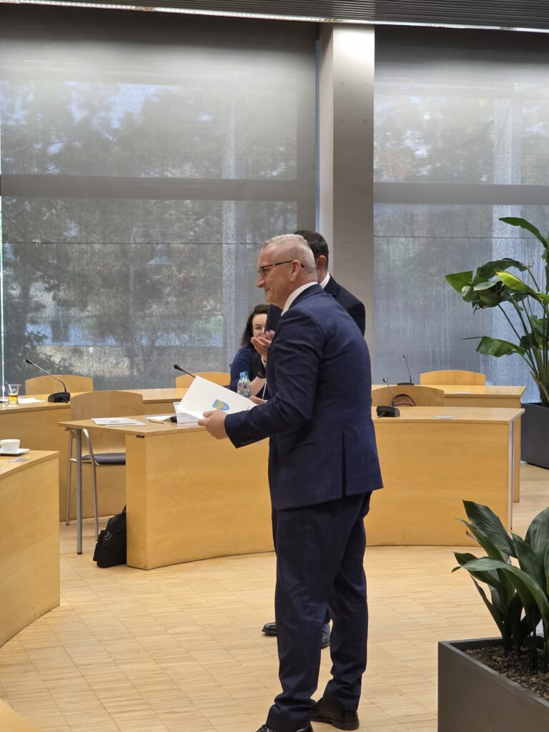 Mężczyzna w ciemnym garniturze i okularach stoi na środku sali konferencyjnej, trzymając w rękach biały dokument z herbem. Został uchwycony z profilu, gdy uśmiecha się w stronę pozostałych osób. W tle widać stoły konferencyjne oraz dużą roślinę doniczkową.