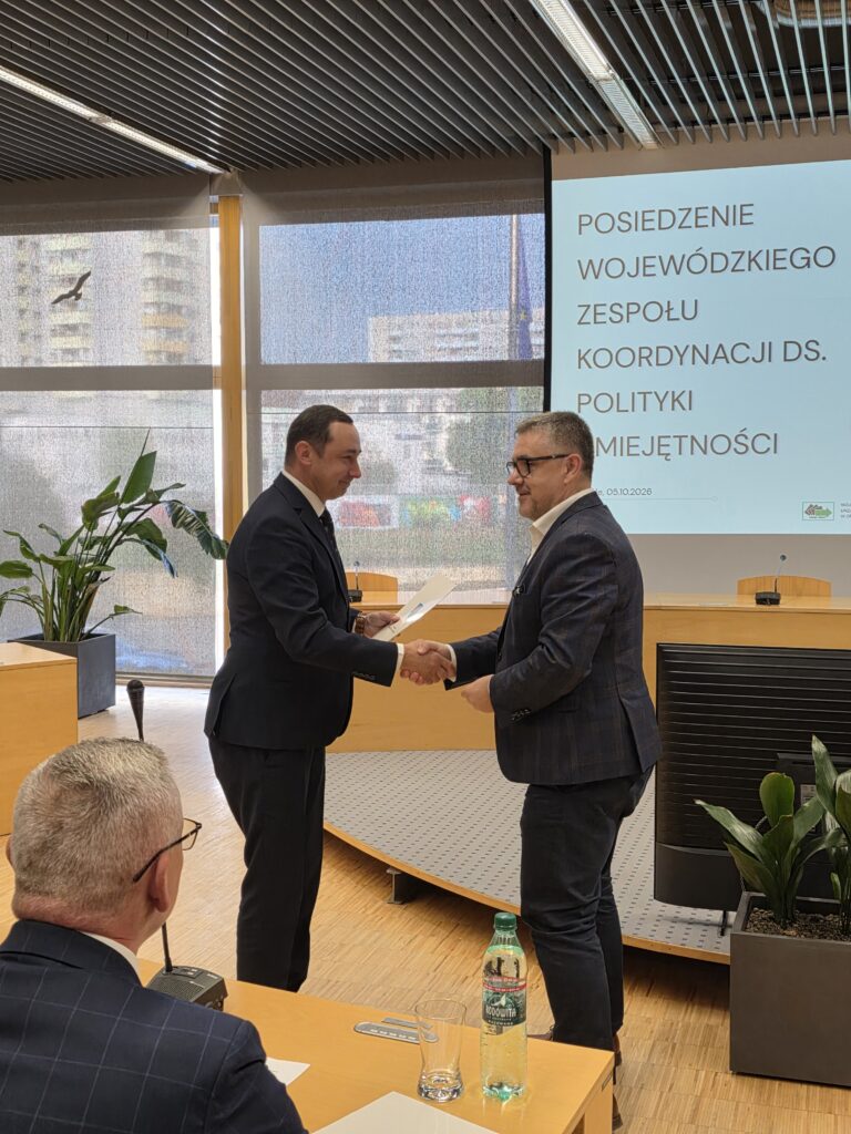 Dwaj mężczyźni stoją naprzeciwko siebie w sali konferencyjnej i podają sobie ręce. Mężczyzna po lewej w garniturze wręcza oficjalny dokument drugiemu mężczyźnie w okularach i marynarce w kratę. Po prawej stronie widać ekran projekcyjny z napisem „Posiedzenie Wojewódzkiego Zespołu Koordynacji ds. Polityki Umiejętności”.