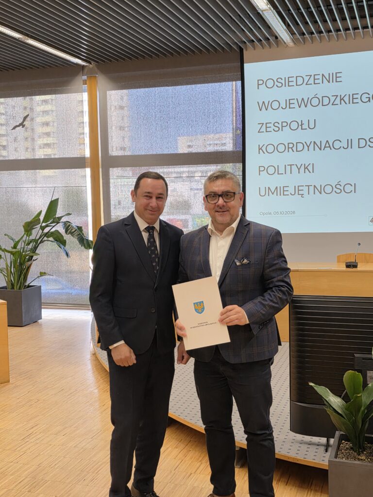 Dwaj mężczyźni w garniturach pozują do zdjęcia, stojąc obok siebie w sali konferencyjnej. Mężczyzna po prawej, w okularach i marynarce w kratę, trzyma w rękach białą teczkę z herbem. W tle widoczny jest ekran z tytułem posiedzenia oraz data 05.10.2026.