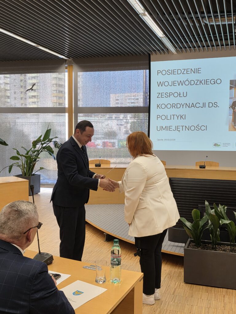 Mężczyzna w ciemnym garniturze podaje rękę kobiecie w jasnym żakiecie i wręcza jej dokument. Kobieta stoi tyłem do obiektywu, lekko pochylona w geście podziękowania. W tle znajduje się ekran z prelekcją oraz oświetlone okna sali.